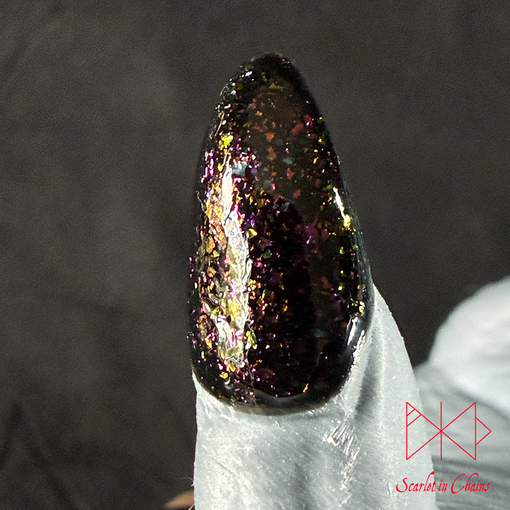 Chaos Witch Nail Varnish Scarlet in Chains and Radioactive Unicorn Collaboration, A close-up of a glossy stiletto-shaped nail tip painted with a dark, iridescent glitter polish that shifts between black, purple, and gold, resting on a white textured surface.