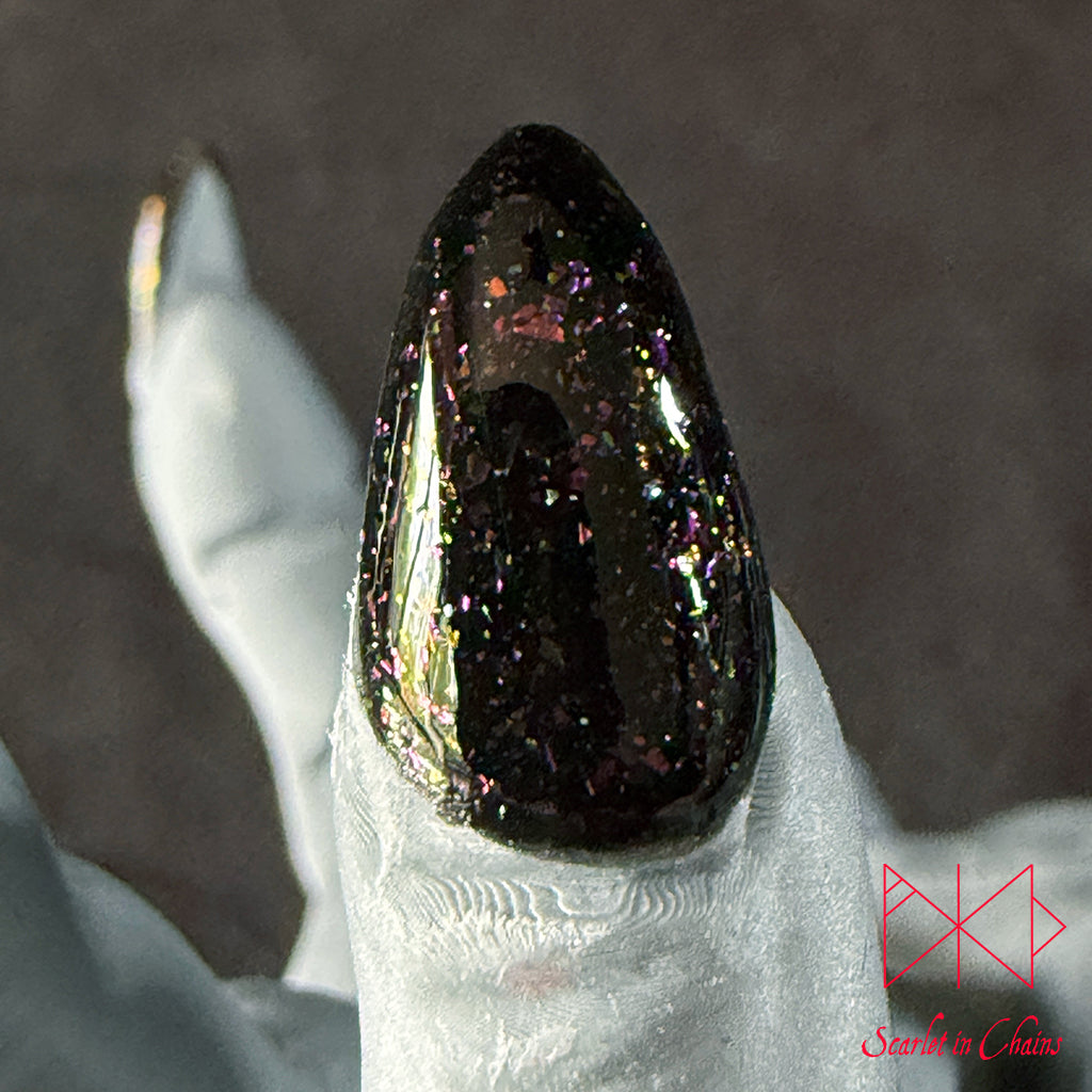 Chaos Witch Nail Varnish Scarlet in Chains and Radioactive Unicorn Collaboration, A close-up of a glossy black stiletto-shaped nail tip decorated with dense iridescent glitter, held on a white gloved finger against a dark grey background.