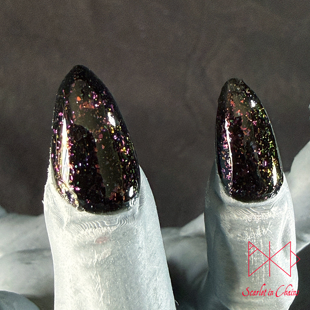 Chaos Witch Nail Varnish Scarlet in Chains and Radioactive Unicorn Collaboration, Two long, glossy stiletto-shaped nails filled with dark glitter polish, displayed on white-tipped fingers against a dark grey background.