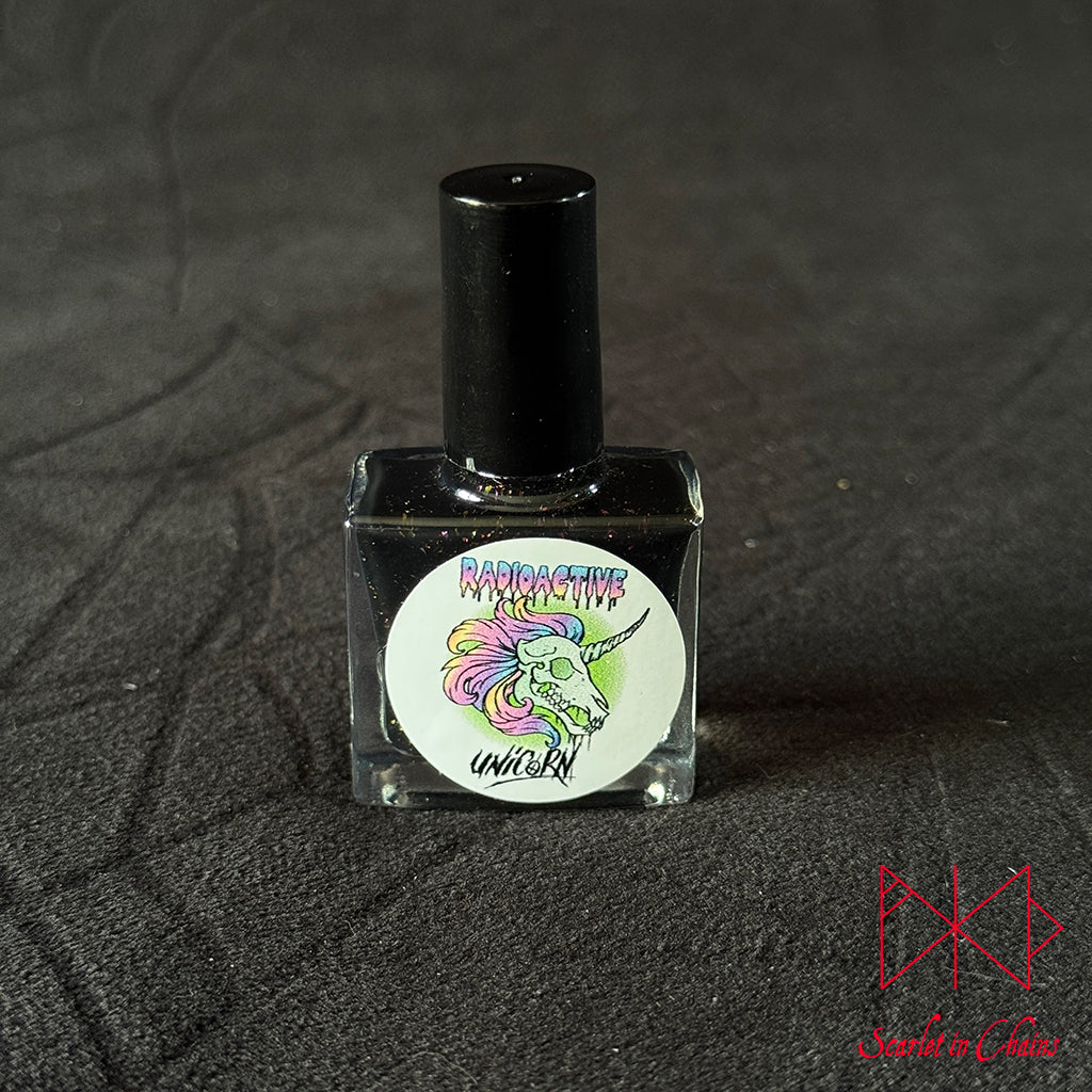 Chaos Witch Nail Varnish Scarlet in Chains and Radioactive Unicorn Collaboration, A square glass bottle of black nail polish with a black cap, featuring a circular white label with a colourful unicorn illustration and the text 'Radioactive Unicorn'. The bottle is set against a dark textured fabric background.