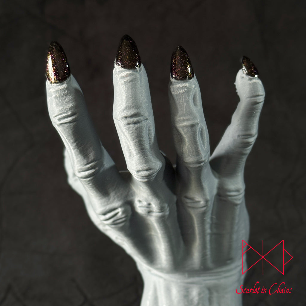 Chaos Witch Nail Varnish Scarlet in Chains and Radioactive Unicorn Collaboration, A white skeletal hand prop with metallic dark Red and Blue glittery fingernails, standing on a white circular base against a dark textured background.