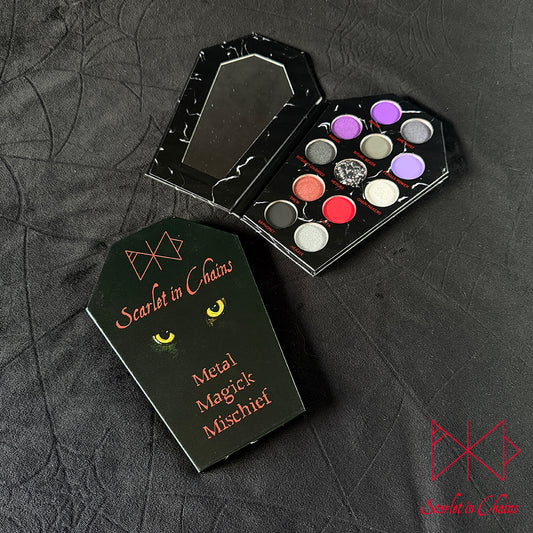 Metal Magic Mischief Makeup Pallet A Scarlet in Chains and Radioactive Unicorn Collaboration. A gothic-themed metal makeup palette set by Scarlet in Chains, featuring a black coffin-shaped case with a mirror and a second closed case displaying the brand name and cat-eye graphic from our Sabra, alongside an open palette showing 12 metallic eyeshadow pans in various colors.