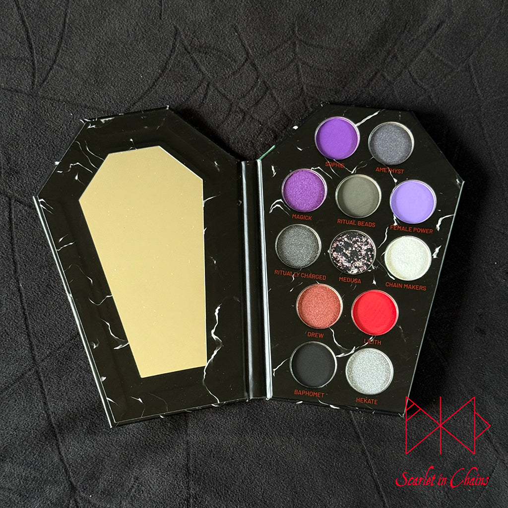 Metal Magic Mischief Makeup Pallet A Scarlet in Chains and Radioactive Unicorn Collaboration. An open gothic coffin-shaped makeup palette with a mirror on one side and 12 eyeshadow pans in various colorus on the other, set against a black textured fabric background.