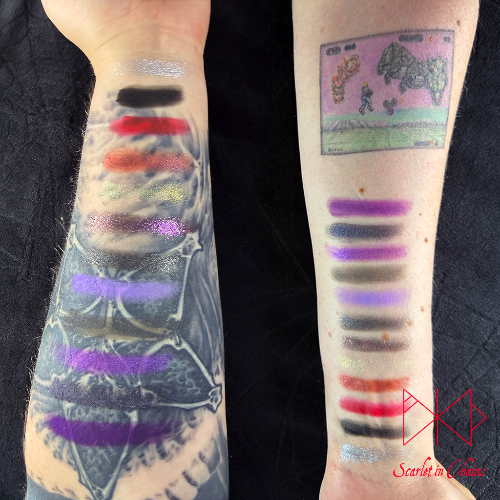 Metal Magic Mischief Makeup Pallet A Scarlet in Chains and Radioactive Unicorn Collaboration. Our two tattooed forearms displayed side by side, each with a vertical row of eyeshadow colour swatches applied on the skin. The left forearm shows black, red, orange, gold, purple, and silver shades.