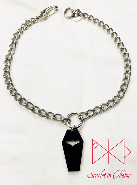 Coffin choker shown flat on a white background. Stainless Steel chain choker with stainless steel O ring at it centre. With a black perspex coffin pendant with a bat cut out suspended from the ring. At the back of the choker a strong easy to use stainless steel clip and an o ring to fasten it to. completed with a stainless steel hand stamped scarlet in chains logo tag