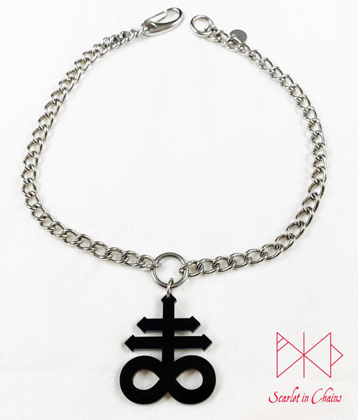 flat shot of Leviathan cross choker on white background. Stainless steel chain choker with stainless steel O ring at centre. Suspended from the O ring is a black perspex Leviathan cross charm. with an easy to use stainless steel clasp and an o ring to close the choker, finished with a hand stamped scarlet in chains logo tag.
